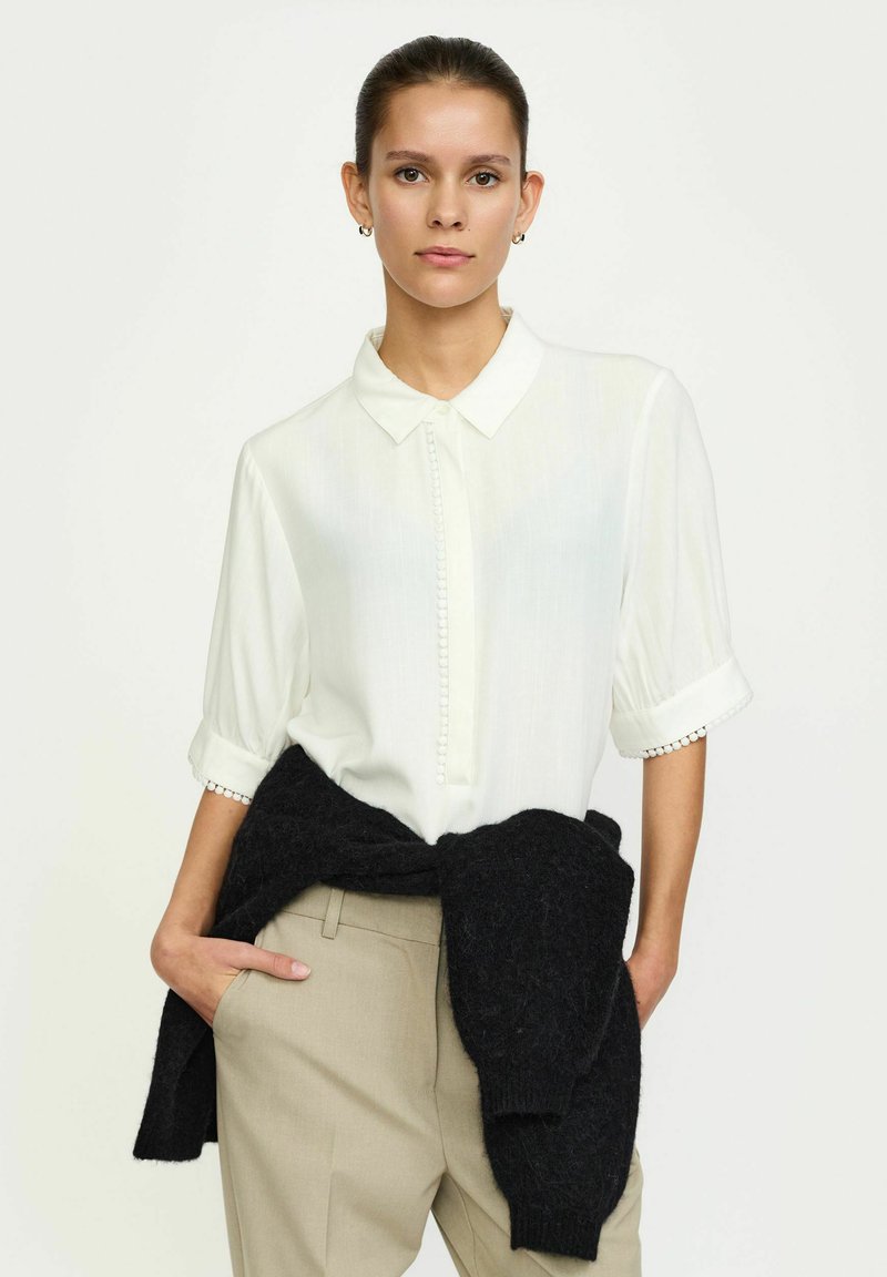 Bijela bluzica kratkih rukava s dugmadi sprijeda i suptilnom vezom na manžetama, uparena s bež hlačama i crnim puloverom vezanim oko struka.