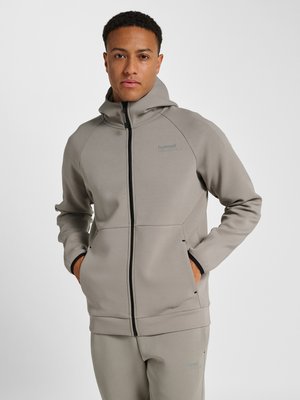 Mand iført beige hoodie med lynlås og matchende joggingbukser med hænderne i lommerne, stående mod en ensfarvet hvid baggrund.