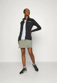 Μαύρο μπουφάν με φερμουάρ, λευκή μπλούζα, ανοιχτό πράσινη φούστα και μαύρα αθλητικά παπούτσια με υφή; περιέχει λογότυπα.