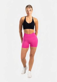 Svart sport-bh och rosa högmidjade shorts i ett mjukt, stretchigt tyg. Vita sneakersdetaljer. Modellen står självsäkert med händerna på höfterna.