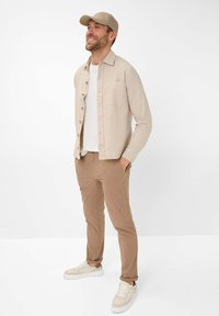 Beige overhemd met knopen over een wit T-shirt, tan broek en lichtgekleurde sneakers. Draagt een bijpassende beige pet en staat tegen een witte achtergrond.
