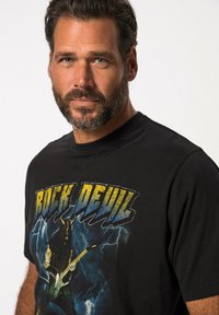 Černé bavlněné tričko s grafickým designem s nápisem "ROCK DEVIL" tučným písmem, zobrazující postavu hrající na elektrickou kytaru.