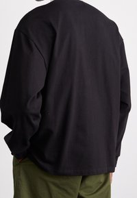 Schwarzes Langarmshirt aus weicher Baumwolle, von hinten gezeigt, mit lockerem Schnitt und abgerundetem Saum, kombiniert mit olivgrünen Hosen.