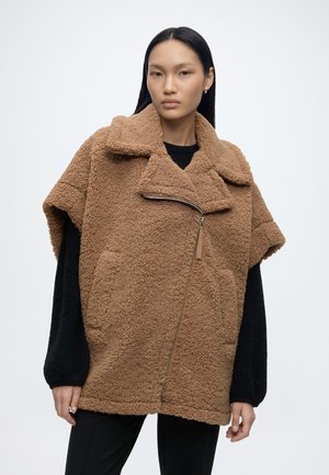 Femme portant une veste marron oversize texturée à fermeture éclair avec un col large, par-dessus un haut à manches longues noires, debout devant un fond clair uni.
