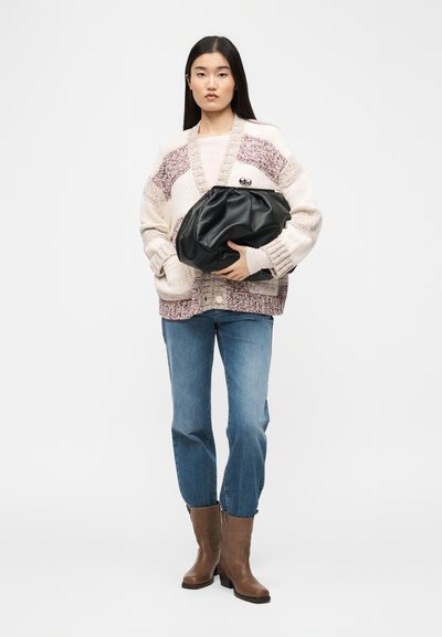 Γυναίκα που φοράει χοντρή πλεκτή ζακέτα, μπλε τζιν, καφέ μποτάκια αστραγάλου, κρατώντας μεγάλο μαύρο δερμάτινο φάκελο με λευκό φόντο.