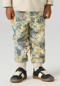 Dziecko w kolorowych spodniach z nadrukiem dinozaurów, czarno-beżowych tenisówkach i białych skarpetkach, stojące na jednolitym tle.