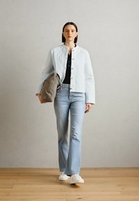 Chaqueta acolchada azul claro con cuello redondo, cierre de botones, combinada con una blusa negra y jeans de campana claros, sosteniendo un bolso gris. Zapatillas blancas.