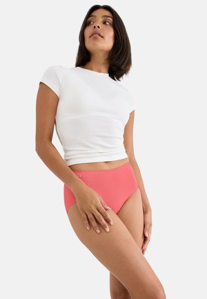 Kvinde iført en hvid tætsiddende kortærmet crop top og koralfarvet højtaljede trusser, stående med den ene hånd hvilende på låret.