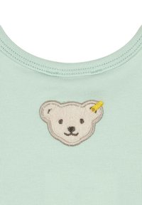 Top in cotone verde menta chiaro con una faccia di orso ricamata e strutturata di colore bianco con un accento giallo, su uno sfondo di tessuto liscio.