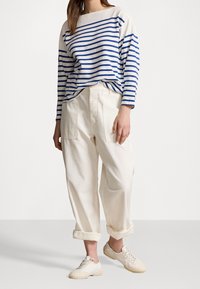 Striped long-sleeve shirt in white and blue, paired with cream-colored loose-fit trousers with side pockets and rolled cuffs, and beige sneakers.