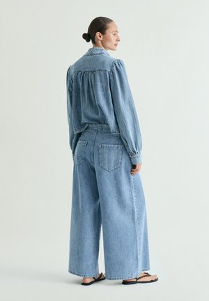 Mujer con el pelo recogido en un moño, vestida con una chaqueta vaquera azul claro y vaqueros de pierna ancha, de pie y mirando hacia un lado sobre un fondo liso.