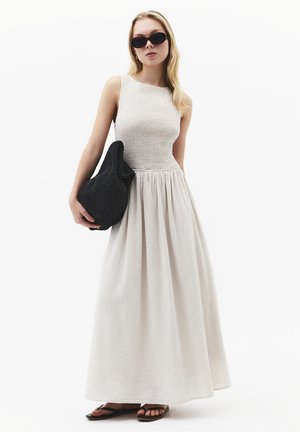 Kobieta w białej sukience maxi bez rękawów, czarnych okularach przeciwsłonecznych, czarnej plecionej torebce na ramię i czarnych sandałach, stojąca na białym tle.