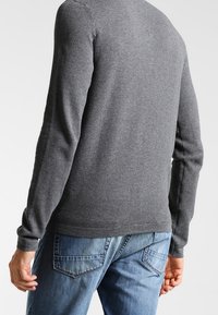 Šedý svetr s dlouhým rukávem s texturovaným pleteným vzorem, pohled zezadu, v kombinaci s modrými džínami. Má kulatý výstřih a žebrované manžety.