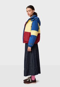 Färgglad dunjacka med blå, gula och röda paneler, med huva, dragkedja och elastiska muddar. Bärs över en lång mörk kjol.