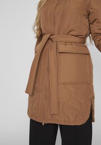Veste matelassée marron avec une ceinture à la taille, équipée d'une fermeture éclair et d'une grande poche plaquée sur le côté. Texture du tissu douce et lisse.