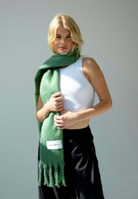 Groene, zachte wollen sjaal met franjes aan de randen, tentoongesteld over een wit shirt; bevat een label met de tekst "ARCTIC FOX" in zwarte letters.