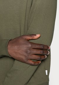 Mörkhyad hand som vilar på den motsatta armen, iförd en långärmad olivgrön tröja med en liten tygetikett nära handleden.