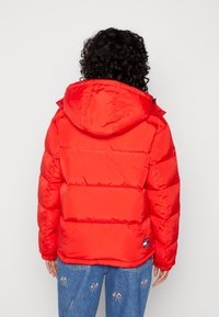 Veste rembourrée rouge avec capuche, présentant un design matelassé et des poignets élastiques. Portée avec un jean en denim brodé, vue de dos.