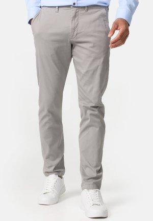 Mand iført lysegrå slim-fit bukser, lyseblå skjorte og hvide sneakers, hænderne i lommerne, stående mod hvid baggrund.