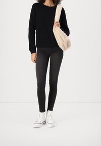 Suéter negro texturizado, vaqueros negros ajustados y zapatillas altas blancas. Bolsa de felpa de color claro colgada de un hombro.