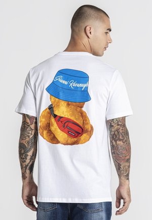 Homme avec des tatouages sur les bras portant un t-shirt blanc avec un graphisme d'ours en peluche portant un chapeau bleu bob et un sac banane rouge dans le dos.