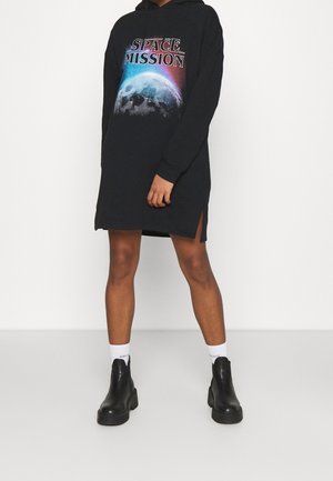 Personne portant une robe à capuche noire avec l'inscription « SPACE MISSION » et un motif de lune, associée à des chaussettes blanches et des bottines noires.