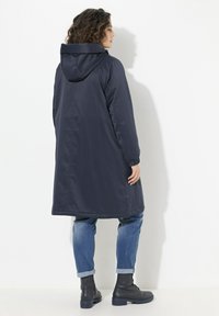 Marinblå lång kappa med huva, tillverkad av slätt tyg. Har elastiska manschetter och rak skärning, kombinerad med ljusblå jeans med uppvikt ärm och svarta stövlar.