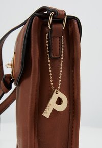 Borsa a tracolla in pelle marrone con catena dorata e grande ciondolo con la lettera "P" dorata attaccato vicino al loop superiore della tracolla.