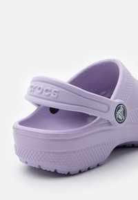 Crocs di colore viola chiaro con superficie testurizzata, schiena aperta e una cinghia. Presentano un logo circolare sul lato e una suola rialzata per una migliore aderenza.