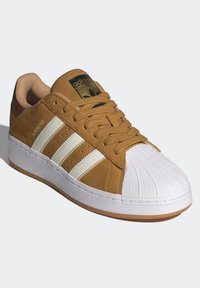 Bruna mockasin med vit gummitå, tre vita läderremsor och en tan innerfoder; har en gul Adidas-logotyp på tungan.