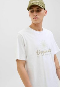 Camiseta blanca de algodón con la palabra "Originals" en estampado dorado, de mangas cortas y corte relajado; combinada con una gorra verde oliva a juego.