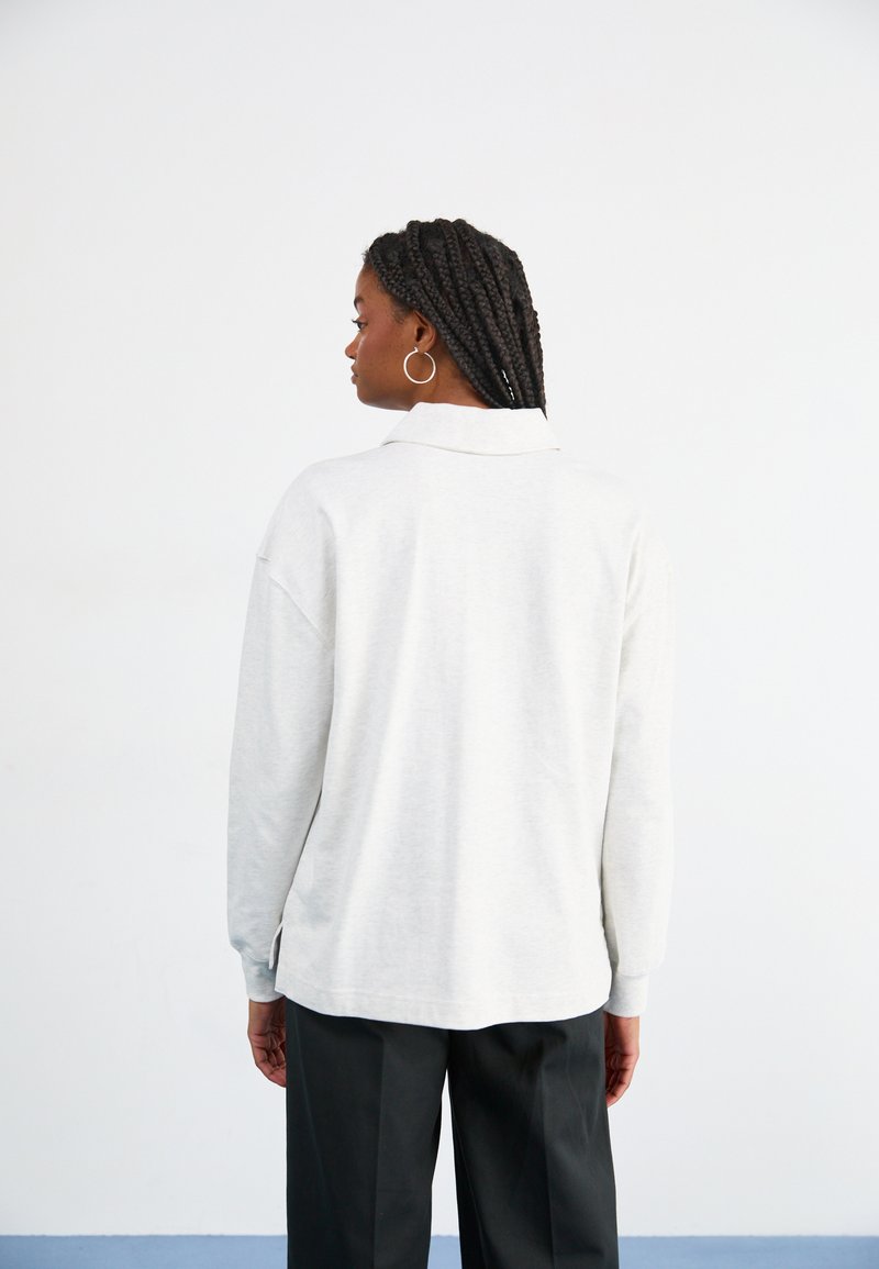 Polo shirt gris clair à manches longues avec col, coupe décontractée, fentes sur les côtés et poignets côtelés, associé à un pantalon noir.