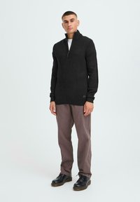 Black knit sweater with a zippered collar, paired with light brown trousers and black footwear. Smooth texture, fitted design.