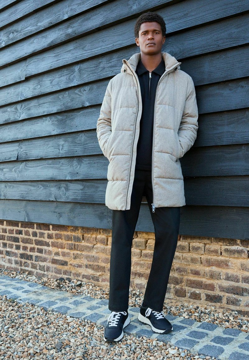 Abrigo acolchado beige con capucha, cierre de cremallera y bolsillos laterales, combinado con pantalones negros y zapatillas deportivas negras, frente a una pared de madera.