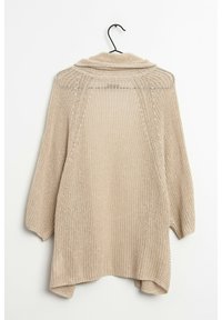 Beige stickad cardigan med en lös, öppen design, med sjalkrage och trekvartslånga ärmar, hängande på en svart krok.