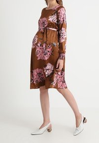 Ruskea kukkamekko, jossa on vaaleanpunaisia ja mustia kuvioita, pitkät hihat ja vyötäröhihna. Yhdistetty valkoisten slingback-kenkien kanssa, joissa on pyöreät korot.