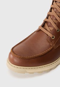 Bota de cuero marrón con cordones color beige, costuras detalladas y suela de goma beige sobre un fondo blanco.