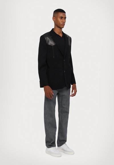 Zwarte blazer met leren schouderaccenten, twee voorkruiken en een knoopsluiting, gecombineerd met grijze jeans en witte sneakers.