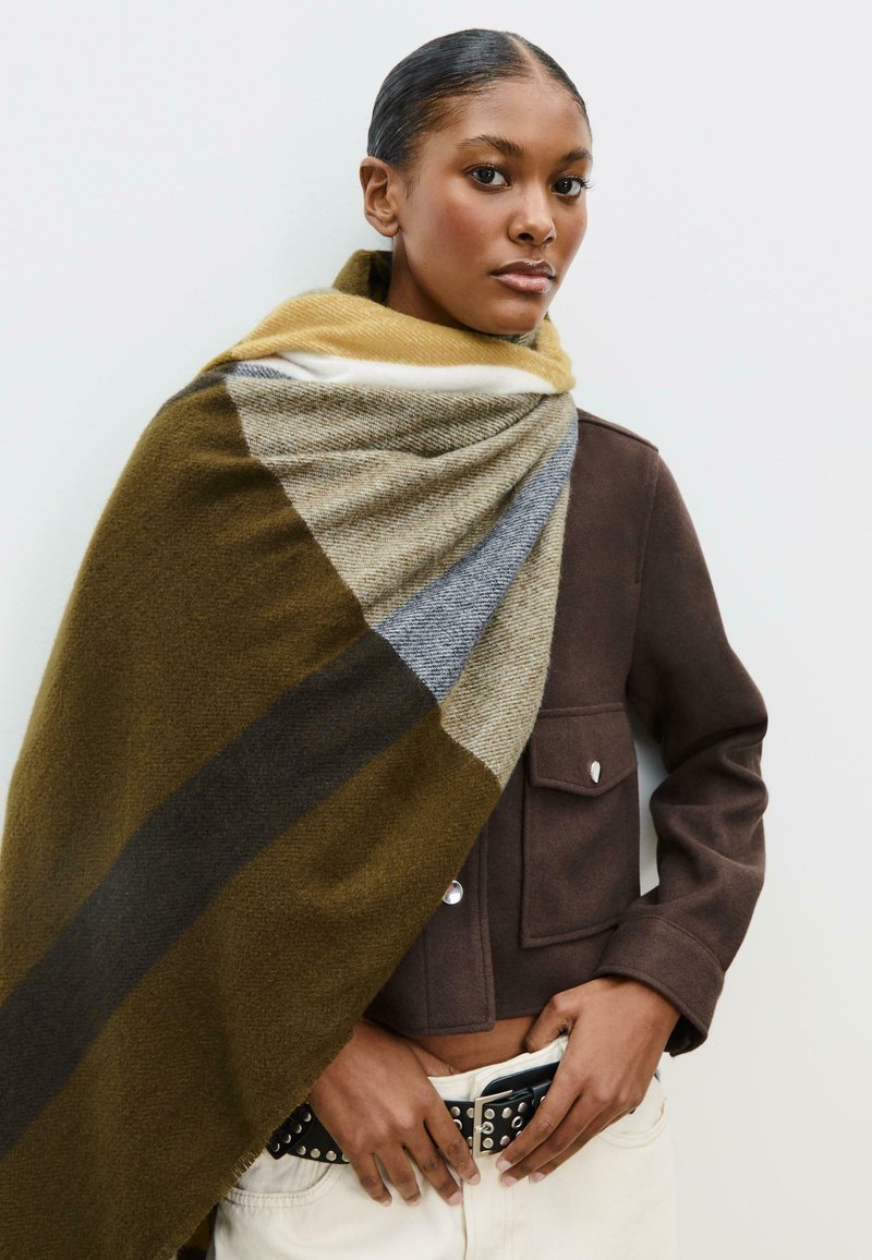 Écharpe tricotée aux motifs géométriques de vert olive, gris et moutarde ; texture douce drapée sur une veste marron. Bords à franges visibles.