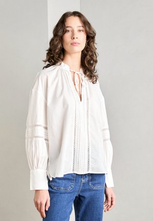 Hvit bluse med knyting ved halsen, blonder detaljer og plisserte elementer. Lange, puffete ermer med tettsittende mansjetter. Kombinert med blå jeans.