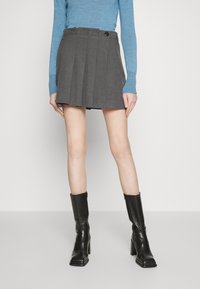 Femme portant une mini-jupe plissée grise, un haut bleu à manches longues et des bottes en cuir noires à talons mi-mollet, debout devant un fond uni.