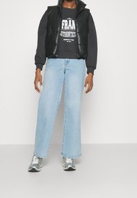 Svart vadderad väst över en grå sweatshirt med vit text, tillsammans med ljusblå vida jeans och grå sneakers. Släta texturer och avslappnad passform.