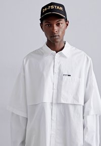 White oversized shirt with a layered design, featuring a chest pocket and button closure. Paired with a black cap displaying gold text.