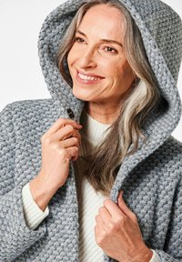 Grau strukturierter Kapuzenmantel mit Knopfverschluss, der ein grob gestricktes Muster und breite Revers aufweist, über einem leichten Pullover getragen.