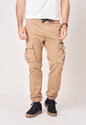 Man draagt beige cargobroek met elastische manchetten en zwarte sneakers, staand tegen een effen lichte achtergrond.