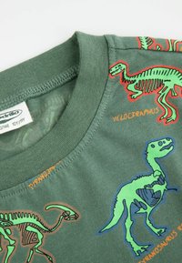 Zielona koszulka z krótkim rękawem z kolorowymi grafikami szkieletów dinozaurów. Na koszulce znajduje się tekst "Wielki Raptor" i "Tyranozaur" w kolorze pomarańczowym.