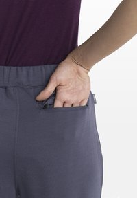 Pantalon de sport gris avec une taille ajustée et une poche zippée. Une main atteint la poche. Le haut est de couleur violet foncé.