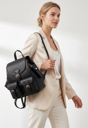 Femme en costume beige portant un sac à dos en cuir noir avec des poches avant bouclées marchant devant un fond clair uni.
