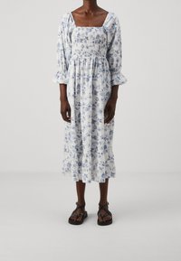 Vit blommig klänning med blå mönster, fyrkantig halsringning, puffärmar och lager av volang i fållen. Bärs med svarta sandaler med remmar.