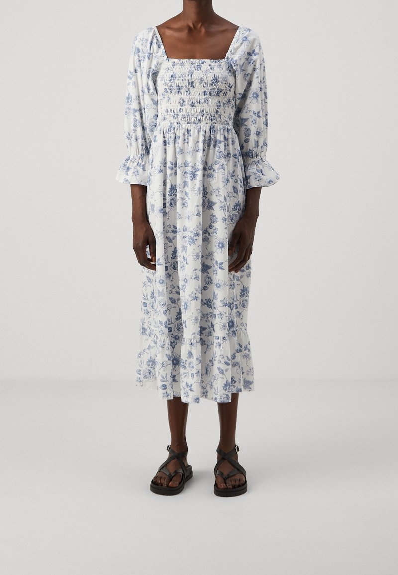 Vit blommig klänning med blå mönster, fyrkantig halsringning, puffärmar och lager av volang i fållen. Bärs med svarta sandaler med remmar.